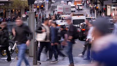 A number of people, blurred by their motion, cross a city street as cars sit nearby.